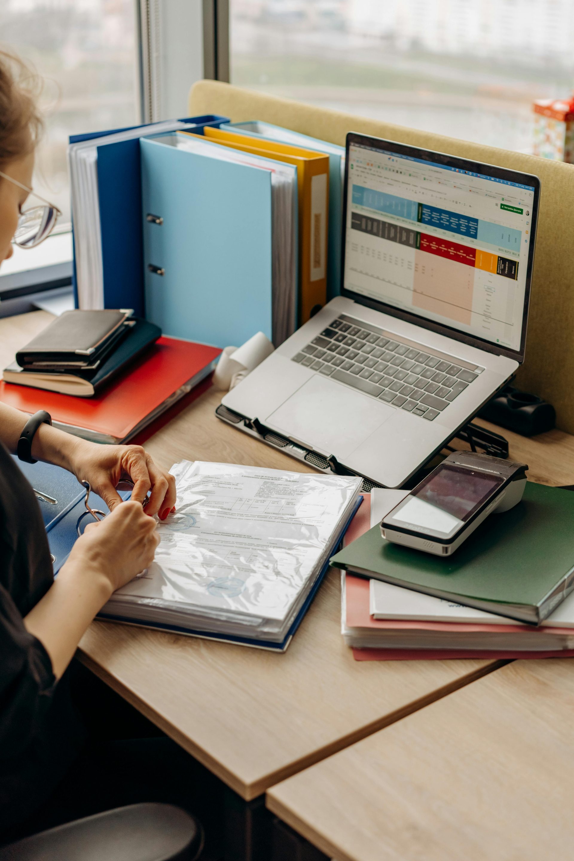 Eine Frau sortiert in einem Büro an einem Schreibtisch mit Ordnern und einem Laptop Dokumente.
